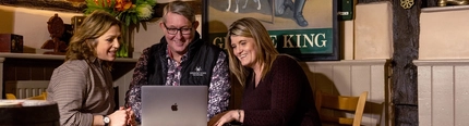 Two women sit on either side of a man as they look at a laptop on the table smiling 