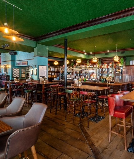 The bar and interior restaurant seating area at the King's Head in Bayswater, with green velvet curtains; green, textured wallpaper on the ceiling; dark wood floor and wall panelling; and a TV hanging above the windows.