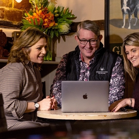 Two women sit on either side of a man as they look at a laptop on the table smiling 