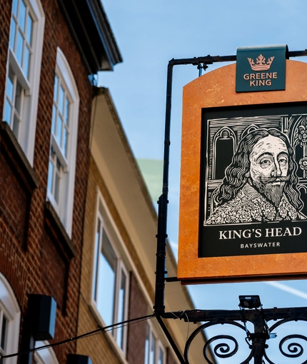 A close up view of the exterior pub sign of the King's Head. The sign features a black and white woodcut-style illustration of a man with a moustache, pointed beard, and shoulder length wavy hair; above the words "King's Head, Bayswater".