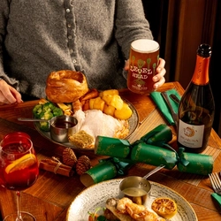 A person is seated at a wooden table. The table is decorated with festive decoration and is  holding a plated turkey dinner.  Only the torso and arm is visible in the image. The person is holding a pint of Level Head.