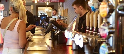 A view looking along the bar at The Flying Horse, featuring customers standing at the bar while members of staff serve them, and a close up view of the beer taps and drinks trays on the bar.
