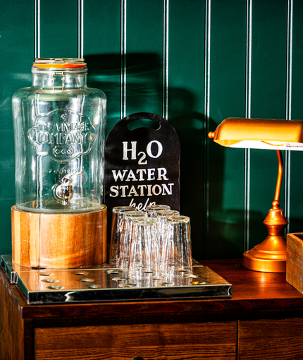 A close up view of the self-serve water station inside The Junction, with a glass water dispenser and upturned highball glasses standing on golden drinks trays, on a wooden cabinet.