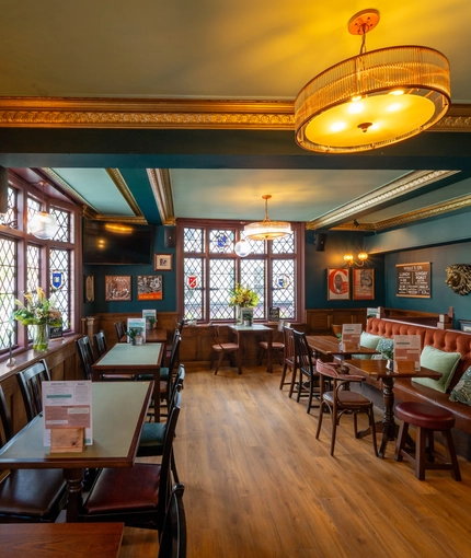 An interior restaurant seating area at The Golden Lion in Soho, with upholstered booth seats, TVs on the walls, golden cornices, and leaded windows with stained glass details.