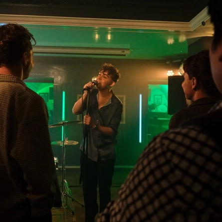 A singer performing on a small pub stage, holding a microphone while audience members watch from the foreground. Green and white illuminated light bars frame the performance area, with a drum kit visible behind the singer. Warm stage lighting and a close audience create an intimate live music atmosphere.
