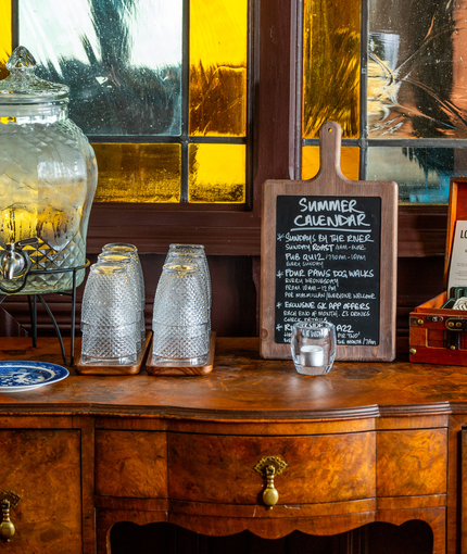 The water station inside The London Apprentice, with glasses and a large glass water dispenser; a small chalkboard sign advertising summer events and deals; and a small briefcase containing coasters and flyers detailing the pub's history; all standing on a wooden sideboard.