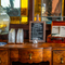 The water station inside The London Apprentice, with glasses and a large glass water dispenser; a small chalkboard sign advertising summer events and deals; and a small briefcase containing coasters and flyers detailing the pub's history; all standing on a wooden sideboard.