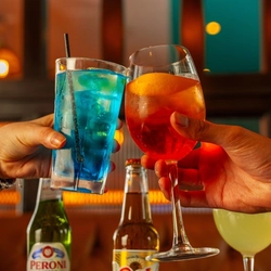 two colourful drinks being held into a 'cheers' position and two bottles of beer Sol and Peroni served in the background at a Flaming Grill venue.