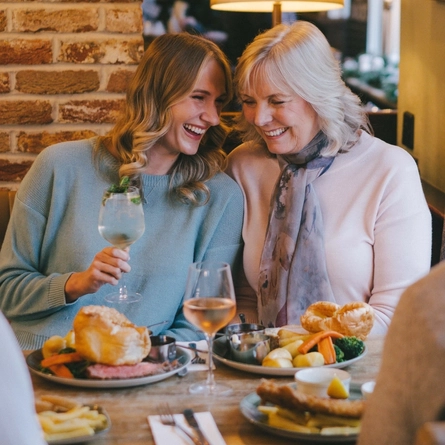 Four people sitting at a restaurant table with plates of food in front of them. One person is holding a cocktail, and a glass of wine also sits on the table.
