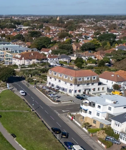 Aerial coastal view of Bournemouth with a wide sandy beach beside blue sea, grassy cliff-top gardens with winding paths, and rows of houses and apartments including The Commodore along the seafront road.