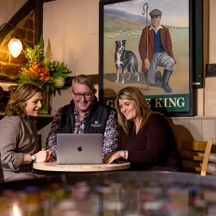 Two women sit on either side of a man as they look at a laptop on the table smiling 