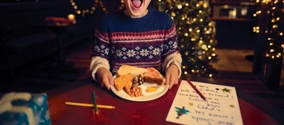A child sitting at a table inside a pub, holding a plate loaded with bread, hash browns, sausages, bacon, baked beans, mushrooms, and a fried egg. A Christmas tree and fairy lights are visible in the background.
