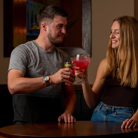 A couple standing at a high table with cocktails in their hands raised in a Cheers type motion.