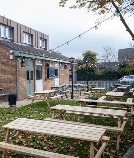The exterior facade, signage, and beer garden seating area at The Longship in Hebburn, with wooden picnic tables, string lights overhead, and a TV on the wall.