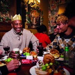 A group of people are gathered around a table enjoying a festive meal. They are wearing paper crowns, and the table is set with plates of food. The background is decorated with Christmas decorations.