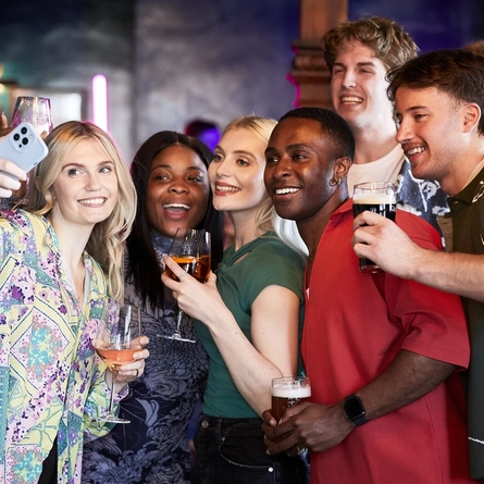 A mixed group of people with drinks in hand, standing, taking a group selfie on a mobile phone.