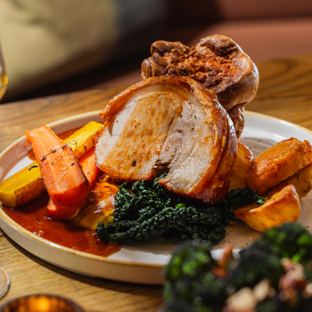 An advertising image showing a plated Roast Pork Loin dish sat on a table within the interior restaurant seating area at The Four Oaks.