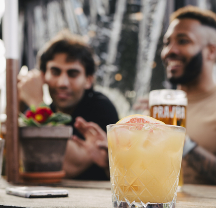 An image focusing on a margarita cocktail sat on a wooden table with a group of friends sat at a wooden table in the background within the exterior beer garden seating area at a Crafted venue.