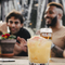 An image focusing on a margarita cocktail sat on a wooden table with a group of friends sat at a wooden table in the background within the exterior beer garden seating area at a Crafted venue.