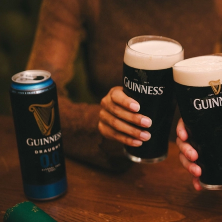 A close up image of 2 friends holding pints of Guinness and celebrating with a cheers within the interior seating area of a Greene King venue.