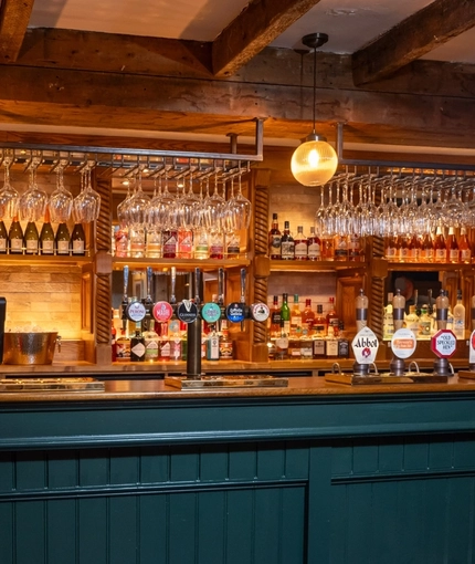 A close up view of the bar inside The Rising Sun in Wistaston, with stone flooring, wooden beams on the ceiling, and wine glasses hanging in racks above the counter.