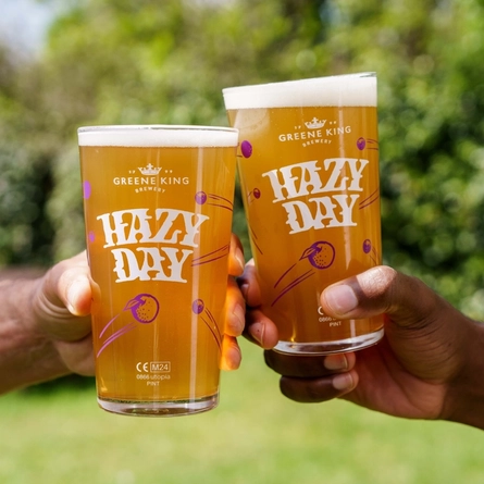 Two hands, each holding a pint of Hazy Day in the air, against a backdrop of greenery in a beer garden.