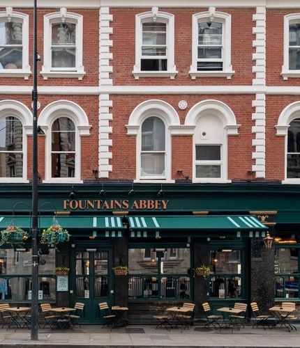 A view from across the road of the exterior facade, signage, and seating area at Fountains Abbey in Paddington.