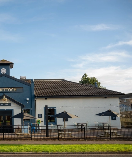 The exterior facade, signage, and beer garden seating area of The Kittoch in East Kilbride, with wooden picnic tables, shade umbrellas, and string lights above the seating area.