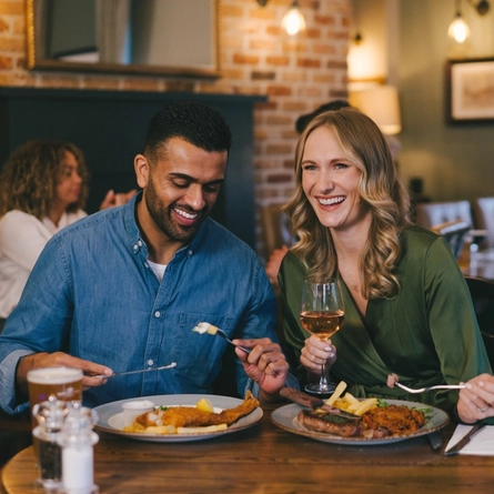 Two people sitting side by side at a small restaurant table, using forks to pick up food from the plates in front of them. One person is also holding a glass of wine.