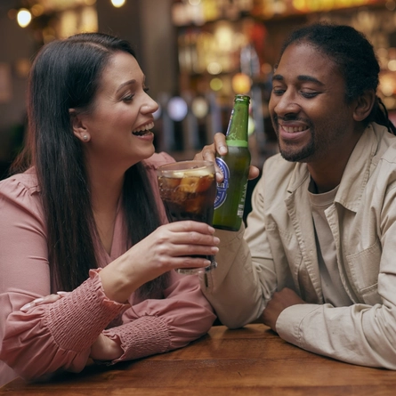 A man holding a bottle of Heineken and a woman holding a glass of Coca Cola, sitting at a table and laughing together.