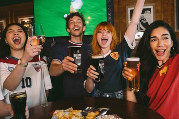 An image showing a group of friends stood at a table watching a football match and enjoying a selection of drinks and snacks while celebrating within the interior seating area at a Greene King venue.