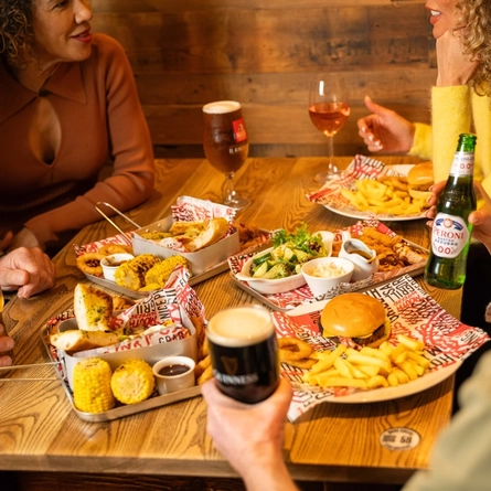 An image showing a group of people sat around a table enjoying mains and sharer dishes with various drinks at a Flaming Grill venue.