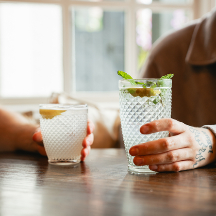 Two people at a wooden table, each holding a cocktail. One cocktail is served in a lowball glass with a slice of lemon and the other is served in a highball glass with lemon and garnished with mint leaves.