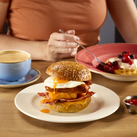 The Big Brekkie Roll at Hungry Horse venues, featuring fried egg, sausage, hash brown and cheese, served on a plate. The plate sits on a table next to a cup of coffee and a bowl containing a waffle topped with berries and cream.