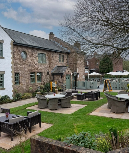 The exterior facade and beer garden seating area at The Malthouse Farm in Whittle-Le-Woods, with tables and wicker chairs on paved sections of the lawn.