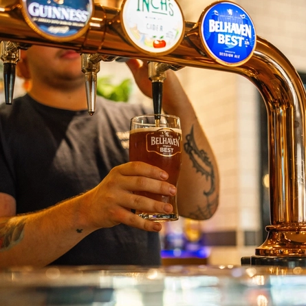 A close up view of a bartender holding a pint glass and filling it with Belhaven Best ale from a beer tap on the bar.