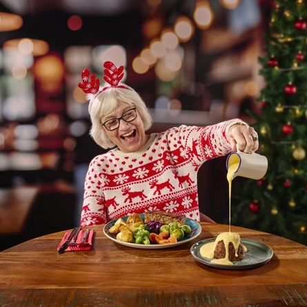 A person wearing reindeer antlers and a Christmas jumper, sitting at a restaurant table and pouring custard over the plate of pudding in front of them. Cutlery, a red serviette, and a plate of Christmas dinner also sit on the table; and a Christmas tree is visible in the background.