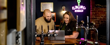 A Man and Woman leaning on the bar looking at a laptop and smiling.