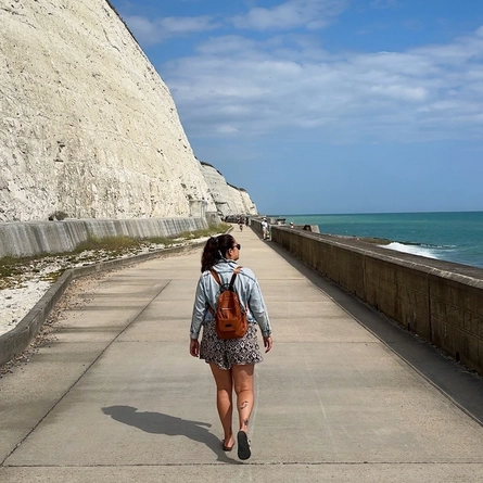An image of a person with her back to the camera, walking along a concrete path with a cliff to her left and the sea to her right.