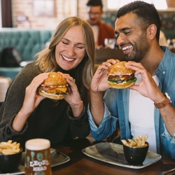 Two people seated at a pub table holding sesame-seed burgers, with portions of chips, a pint of Level Head beer and a glass of white wine.