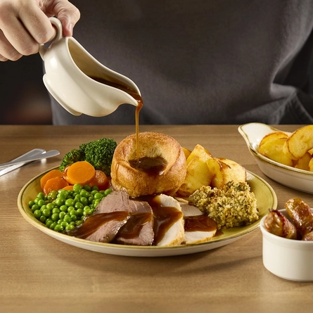 A Duo of Meat roast dinner at Hungry Horse venues, featuring slices of roast meat, roast potatoes, stuffing, Yorkshire pudding, peas, and vegetables, served on a plate. The plate sits on a wooden table next to a ramekin of pigs in blankets and a small roasting dish of roast potatoes, and a person sitting at the table is pouring gravy over the plate from a gravy boat.