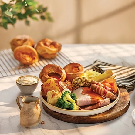 A roast dinner featuring roast meats, Yorkshire pudding, vegetables and gravy served on a plate, and a large slice of chocolate cake served on a plate with a fork. Both plates sit on a table along with a napkin, a jug of gravy, a small pot of sauce, and more Yorkshire puddings in the background.