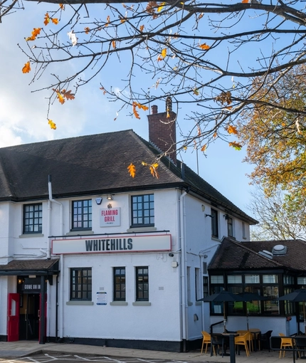 The exterior facade, signage, beer garden seating area, and car park of Whitehills in Northampton, with accessible parking spaces, and shade umbrellas over some of the tables.