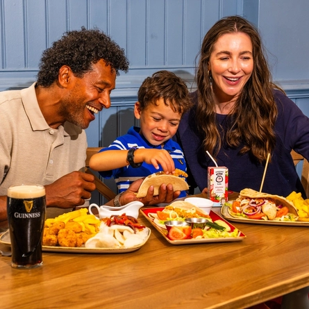 Two adults and a child are seated at a wooden table in a pub, enjoying a midweek meal. The table is filled with dishes including tacos, chips, corn on the cob and breaded bites, alongside a pint of Guinness, a glass of cola with lemon and a children's juice carton. Blue panelled walls and wooden seating create a relaxed pub setting in the background.