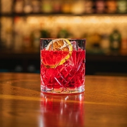 A close up view of an Another Negroni served in a whiskey glass with ice and a dried orange slice. The glass stands on the bar inside a pub.