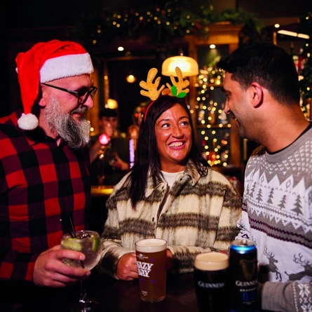 Three people are standing at a tall table with drinks,  in a festive, warmly lit bar. Two of them are wearing Christmas themed headwear.  The background features Christmas decorations, including garlands and lights, as well as hanging glasses above the bar area.