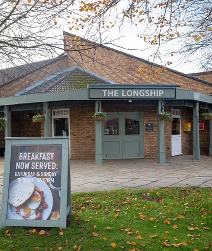 The exterior facade and signage of The Longship in Hebburn, with hanging flower baskets, and a sign board in front of the building which says "Breakfast now served: Saturday & Sunday, 10am - 12pm".