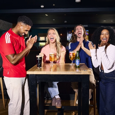 An image showing 4 friends at a table enjoying drinks and celebrating while watching on a football match within the interior seating area at a Flaming Grill venue.