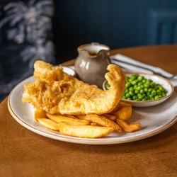 An advertising image of a plated Cod & Chips dish sat on a table within the interior restaurant seating area at a Chef & Brewer venue.