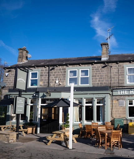 The exterior facade, signage, and seating area of The Horsforth, with wooden picnic tables, shade umbrellas, and string lights above the tables.